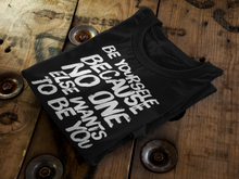 Load image into Gallery viewer, A black t-shirt, folded neatly with its white text visible, sits on a rich wooden table, presenting a blend of elegance and readiness for the wearer&#39;s next escapade.